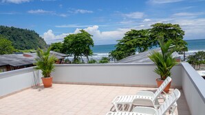 A terrace with white lounge chairs, potted plants, and a view of the ocean and hills. - Mar Arena 27 Apartment (Jacó)