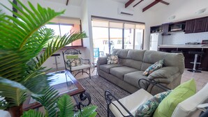A living room with a sofa, coffee table, and chairs, a kitchen area with cabinets and appliances, and large windows allowing natural light. - Mar Arena 27 Apartment (Jacó)