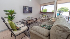 A living room with a sofa, armchair, and coffee table, a TV on the wall, and a large window with a view of trees. - Mar Arena 27 Apartment (Jacó)