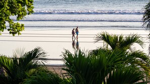 A beach with two people walking, palm trees, and a calm sea. - Mar Arena 27 Apartment (Jacó)