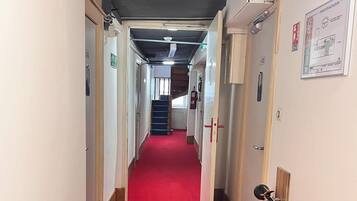 Un couloir recouvert d’une moquette rouge, avec des portes de chaque côté, un extincteur sur le côté droit et un escalier au fond.