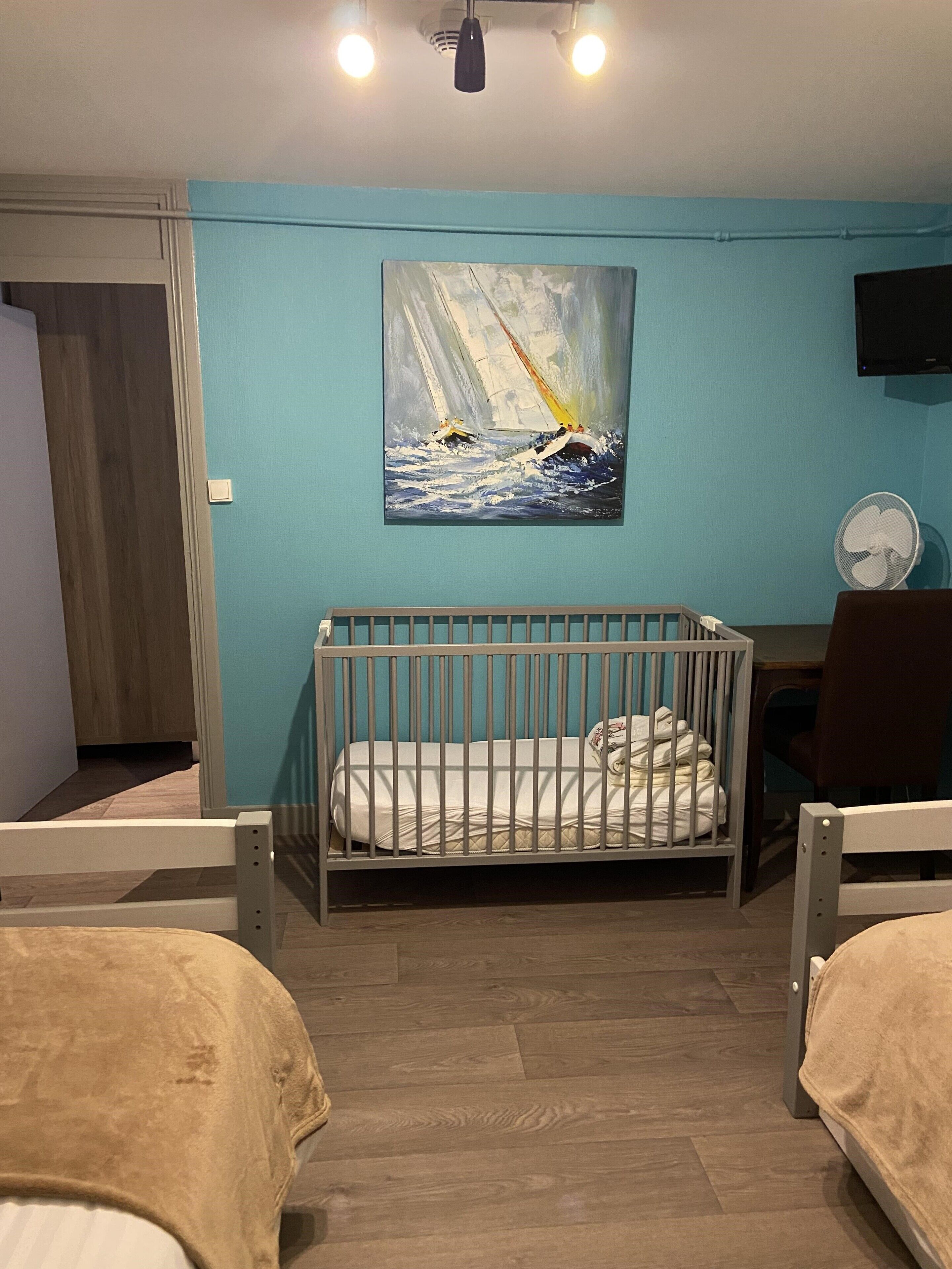 Una habitación con cuna, cama, un cuadro de veleros, televisor y ventilador.