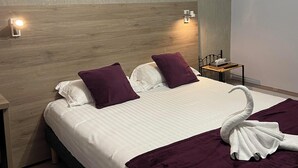 A neatly made bed with white linens and purple pillows, a wooden headboard, a bedside table with a lamp, and a swan-shaped towel on the bed. - Hôtel L'Univers (Chatellerault)