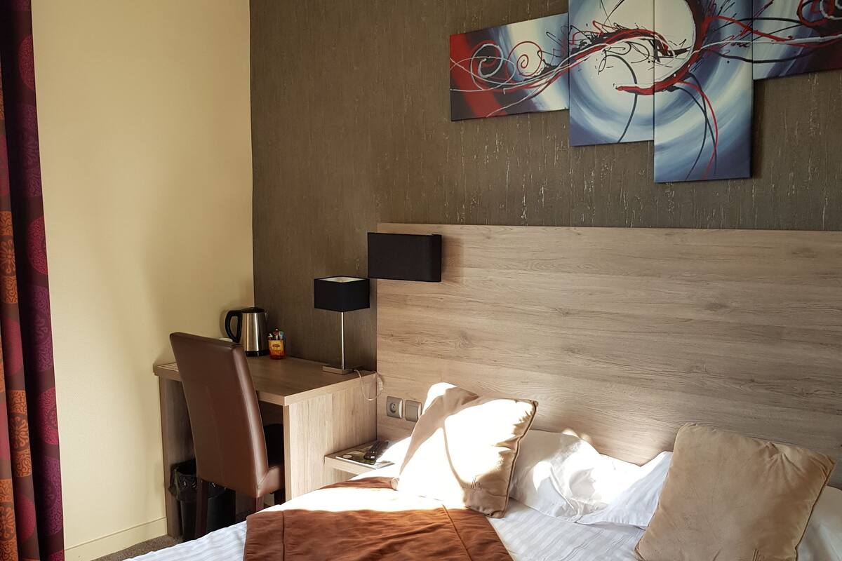 A hotel room with a bed, a desk, a chair, a lamp, and a painting on the wall.