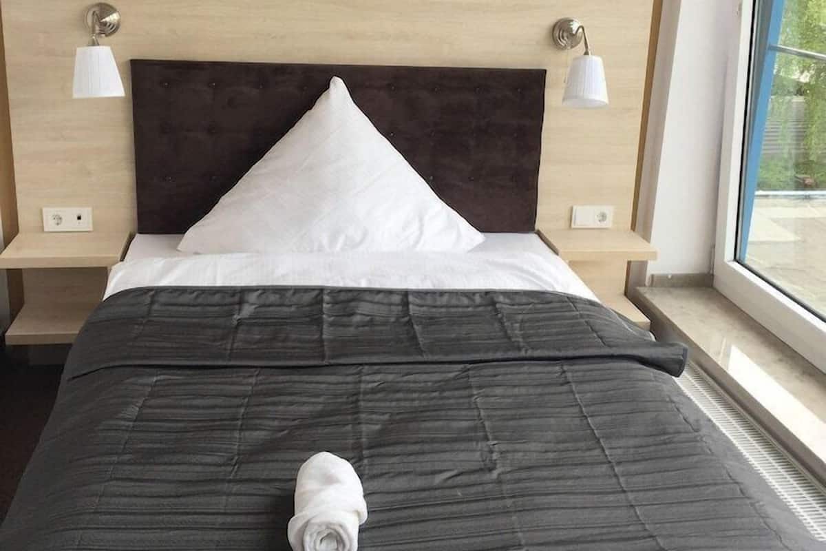 A hotel room with a dark headboard, white pillows, a rolled towel on the bed, two bedside lamps, and a painting on the wall.
