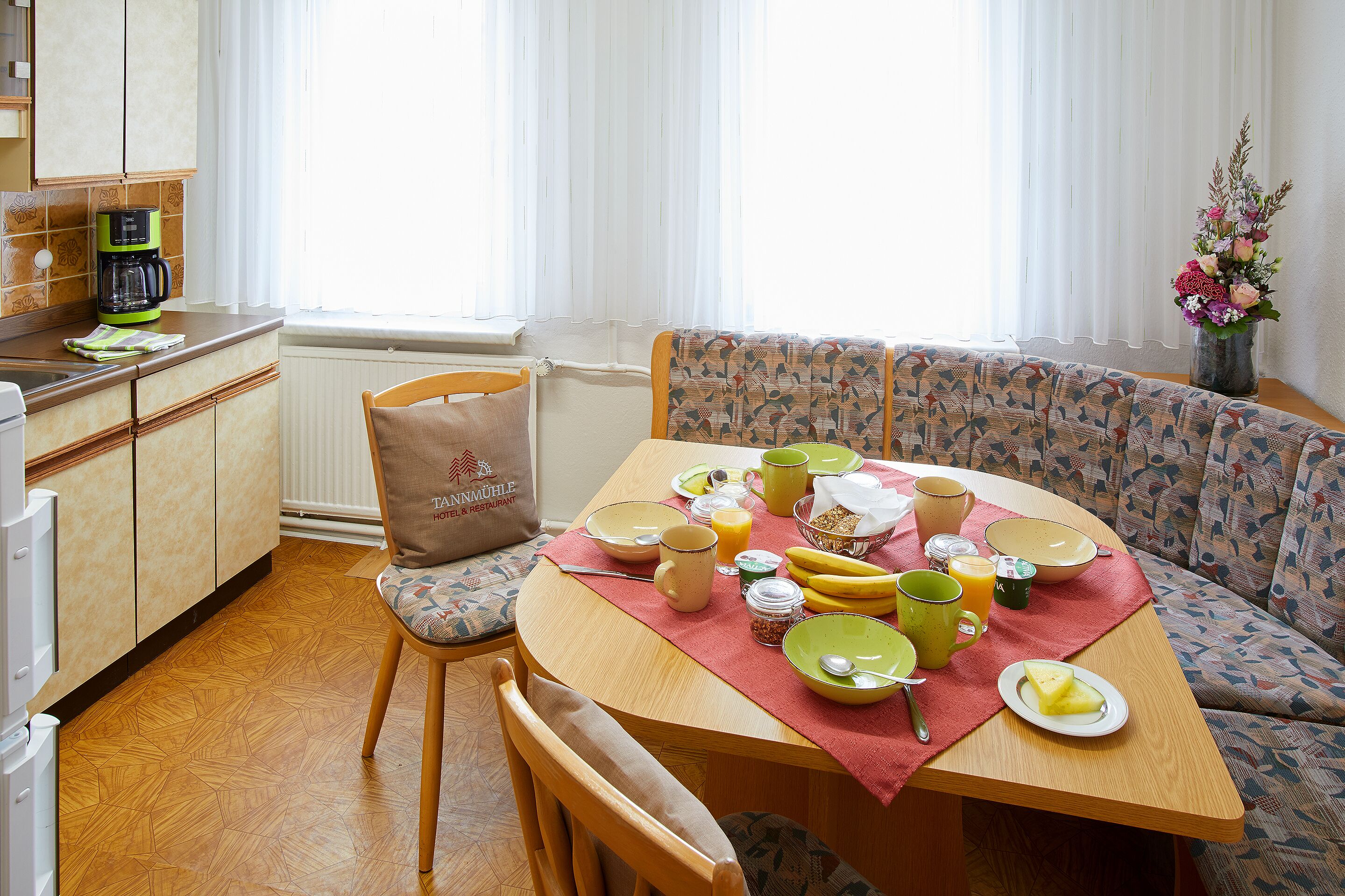 A dining area with a wooden table set for breakfast, a sofa, and a kitchen in the background.