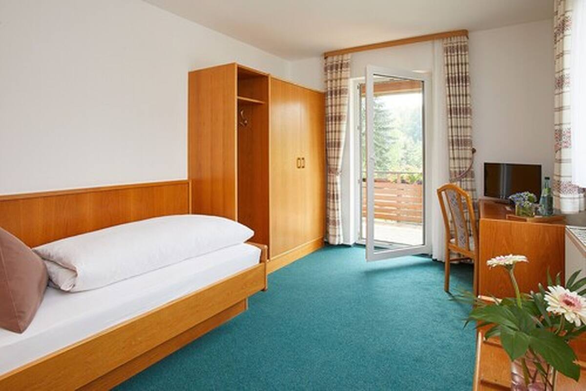 A bedroom with a bed, a wooden wardrobe, a desk with a chair, and a television.