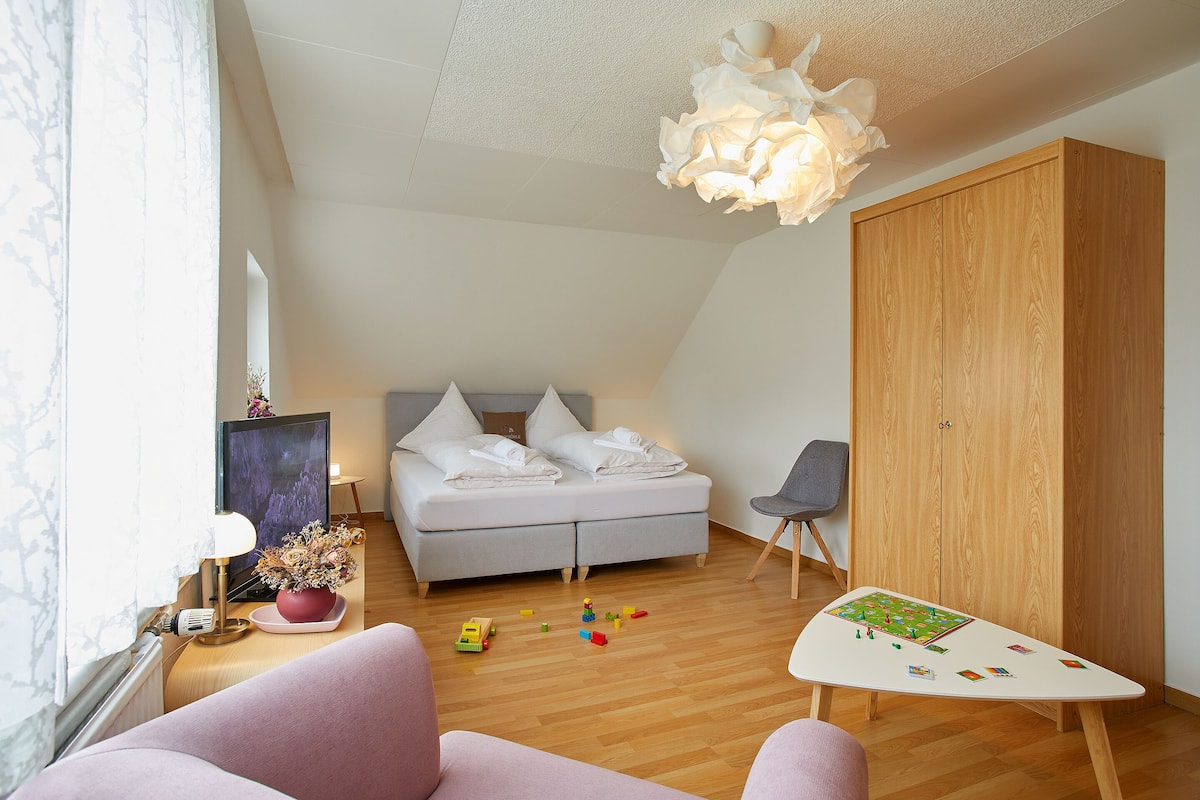 A bedroom with a bed, a wooden wardrobe, a desk with a lamp, and a small table with toys.