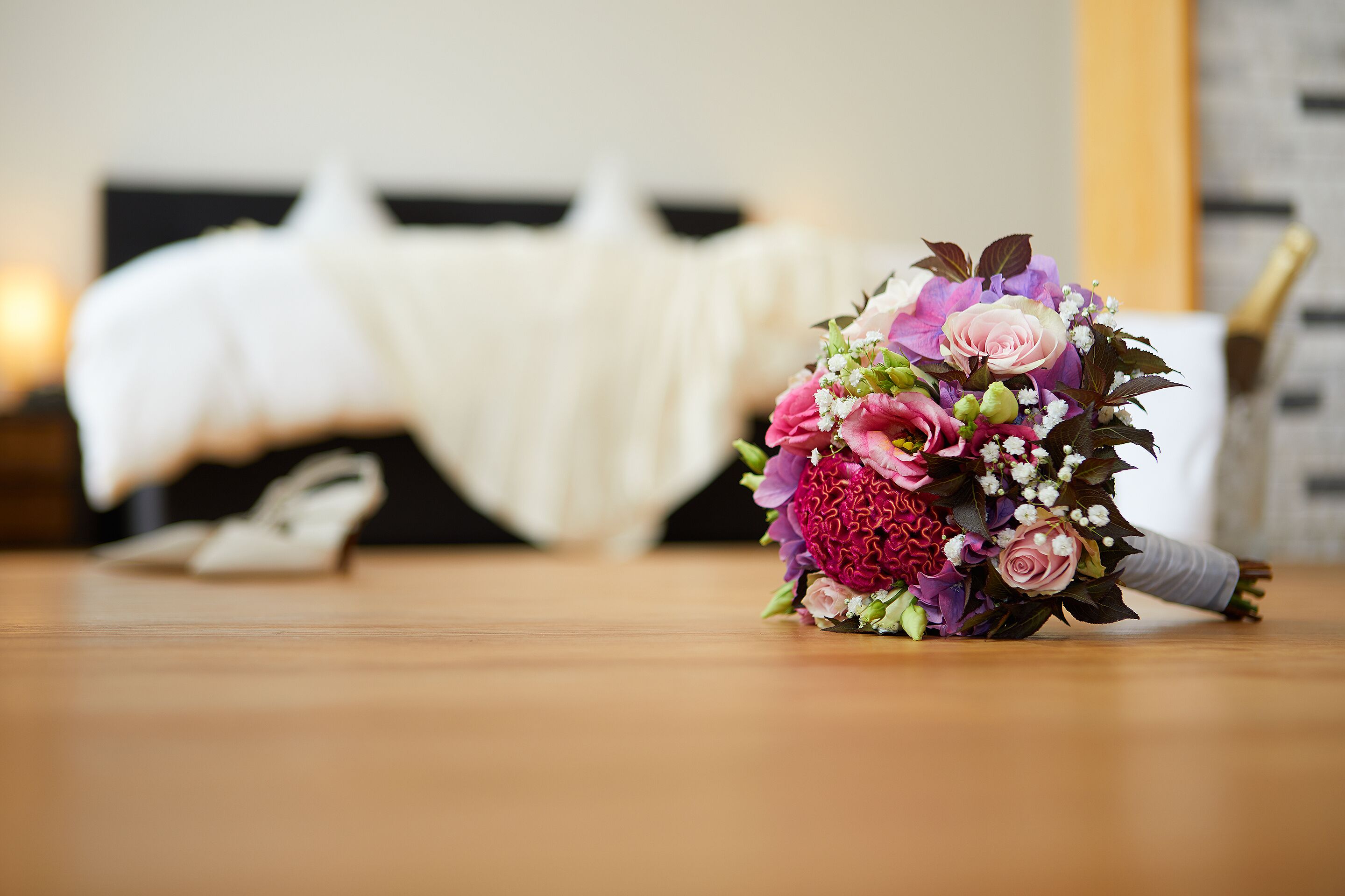 A bouquet of flowers on a wooden table with a bed and a nightstand in the background.