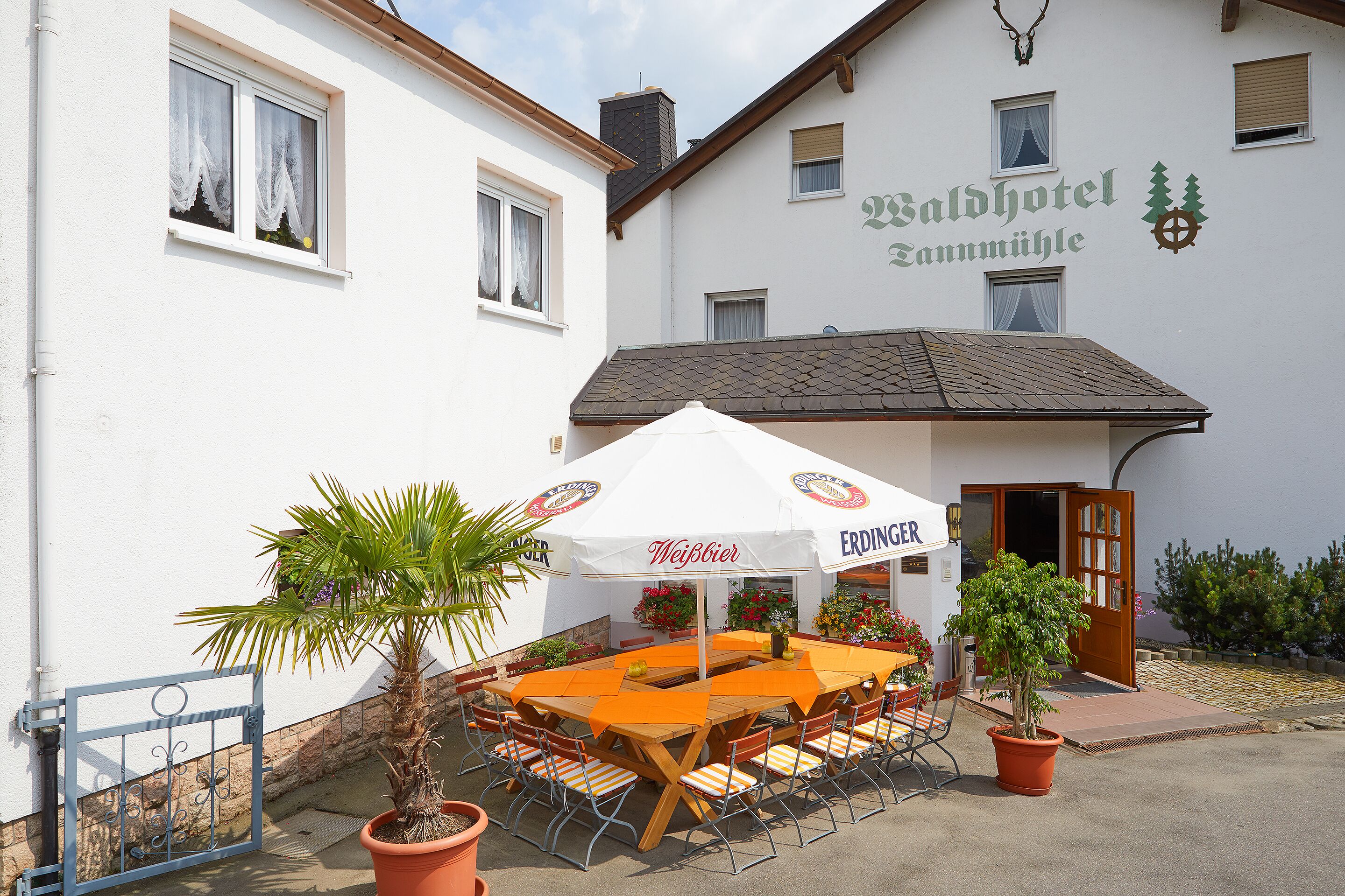 A hotel exterior with a wooden table and chairs under a white umbrella, a potted plant, and a metal gate.