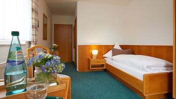 A hotel room with a single bed, a bedside table with a lamp, a bottle of water, and a vase with flowers.