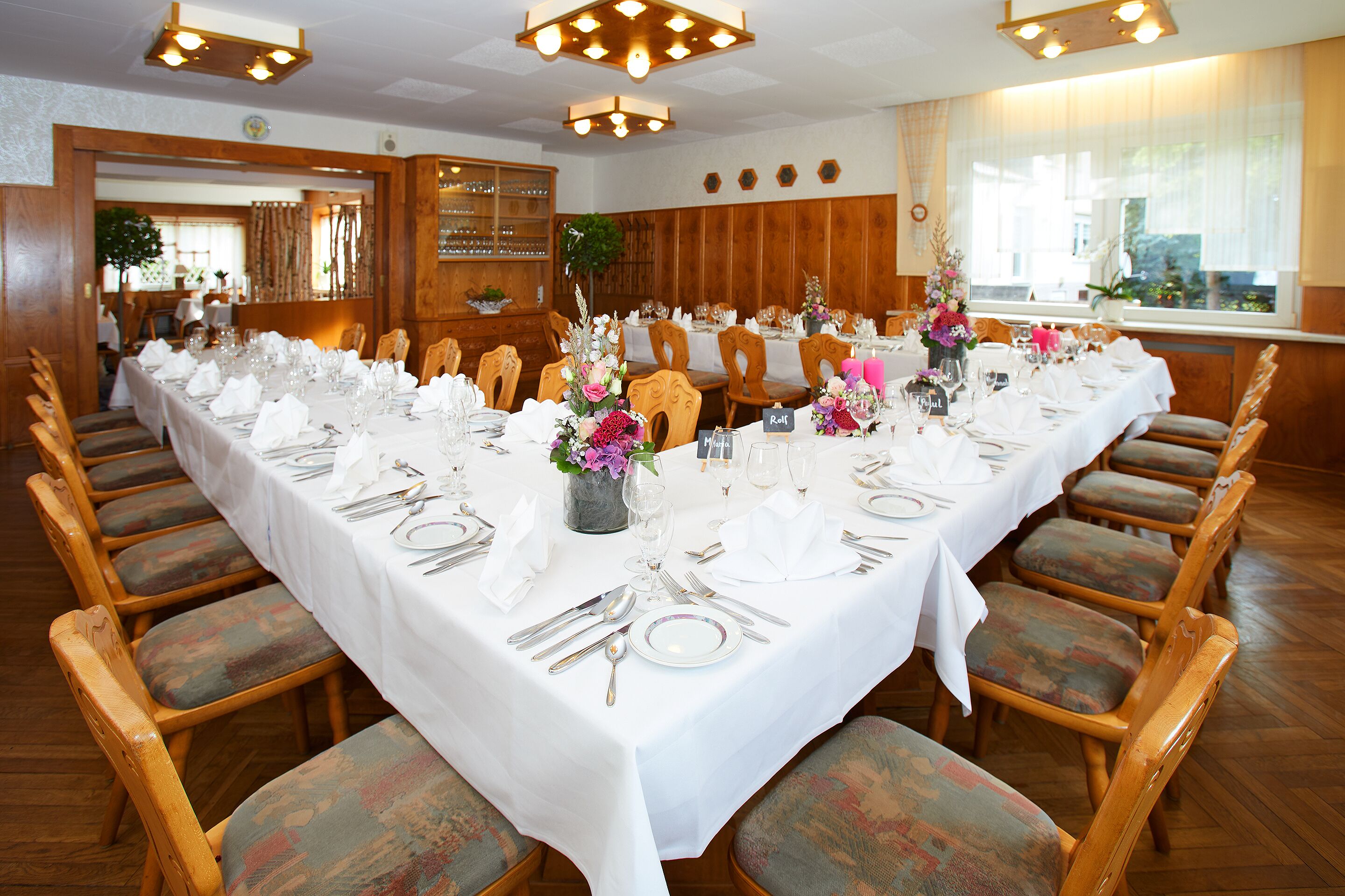 A dining area with a long table set for a meal, wooden chairs, and a wooden panel wall.