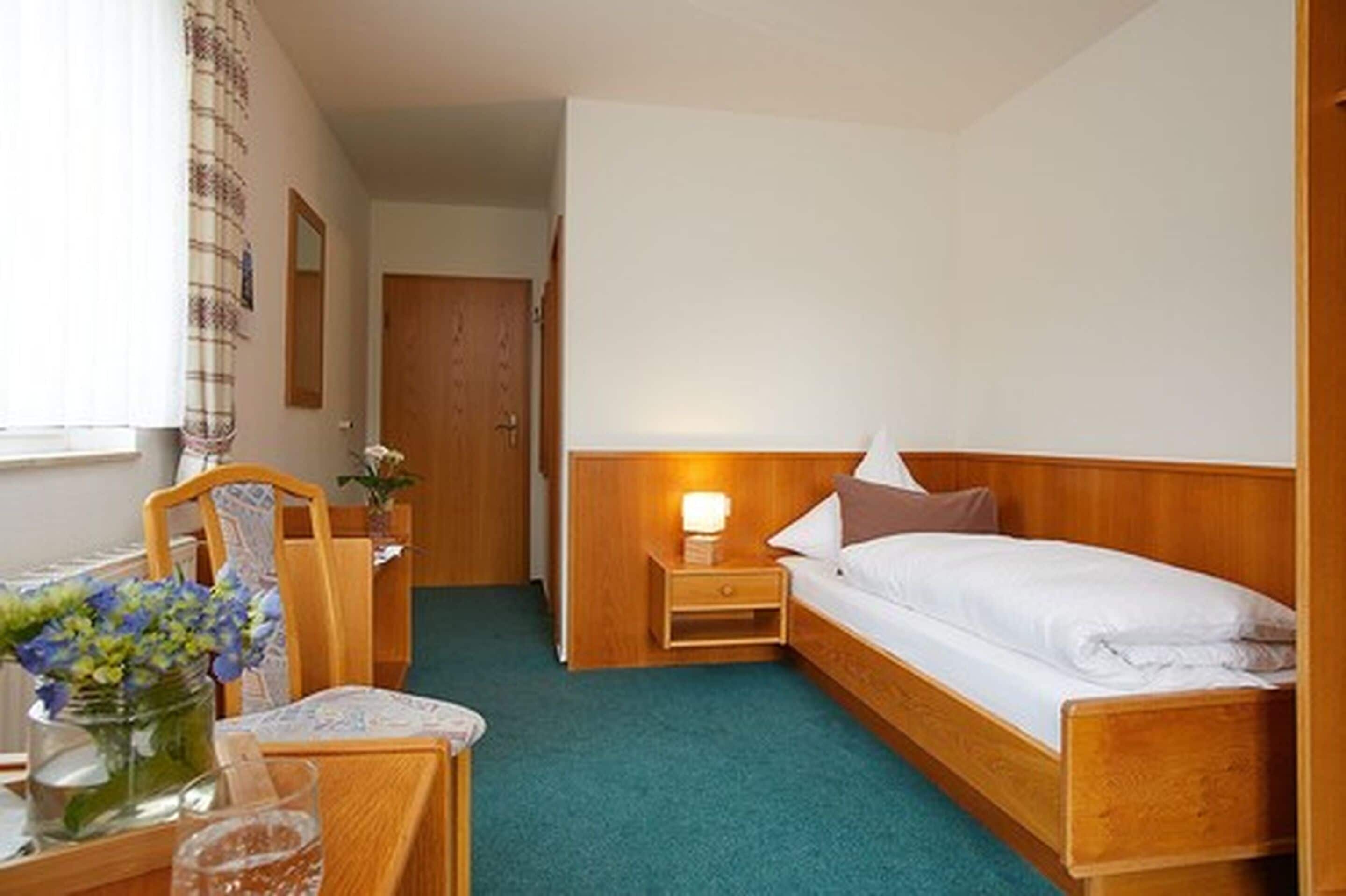 A compact hotel room with a single bed, a bedside table with a lamp, a small table with a vase of flowers, and a chair.
