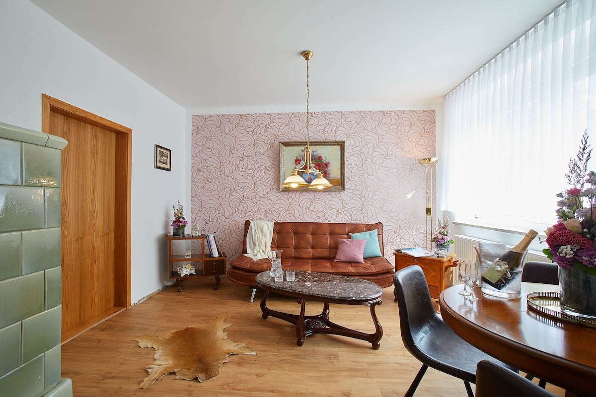 A living room with a wooden floor, a brown leather sofa, a coffee table, and a dining table with chairs. There is a glass of water on the coffee table and a bottle of champagne on the dining table.