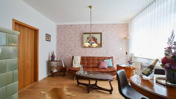 Ein Wohnzimmer mit Holzboden, einem braunen Ledersofa, einem Couchtisch und einem Esstisch mit Stühlen. Auf dem Couchtisch steht ein Glas Wasser und auf dem Esstisch eine Champagnerflasche.