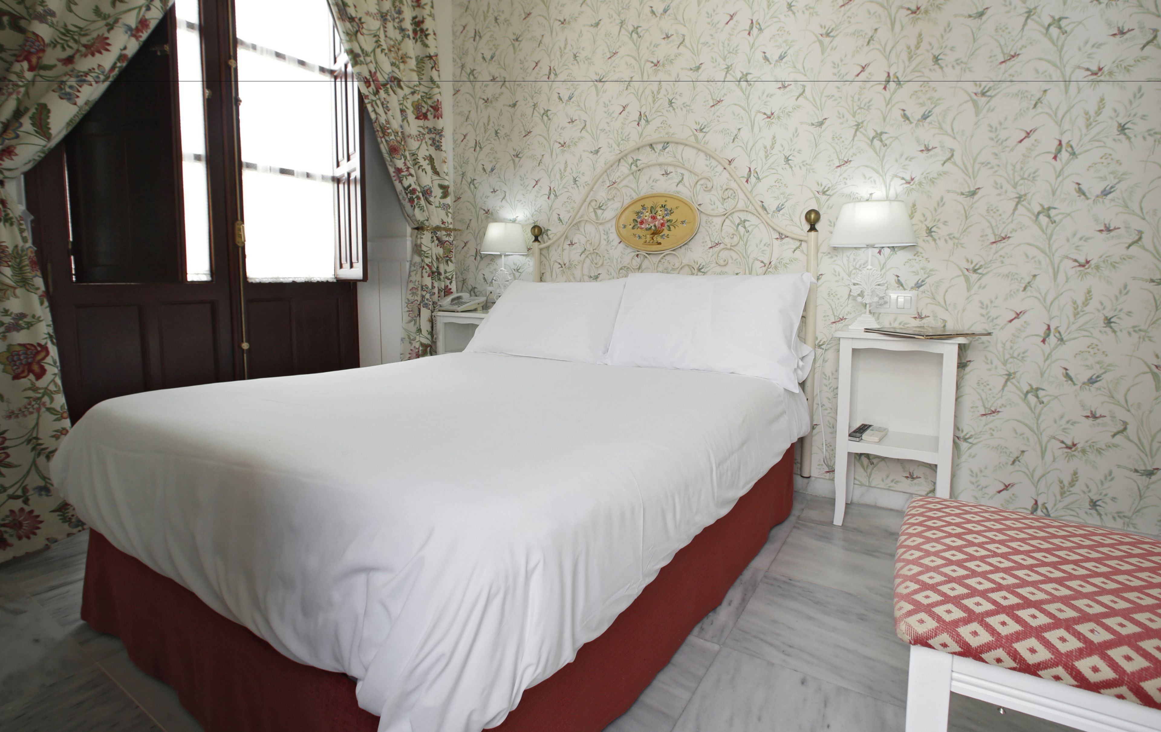 Una cama bien tendida en una habitación con papel tapiz floral, una puerta de madera y una mesita de noche con una lámpara.
