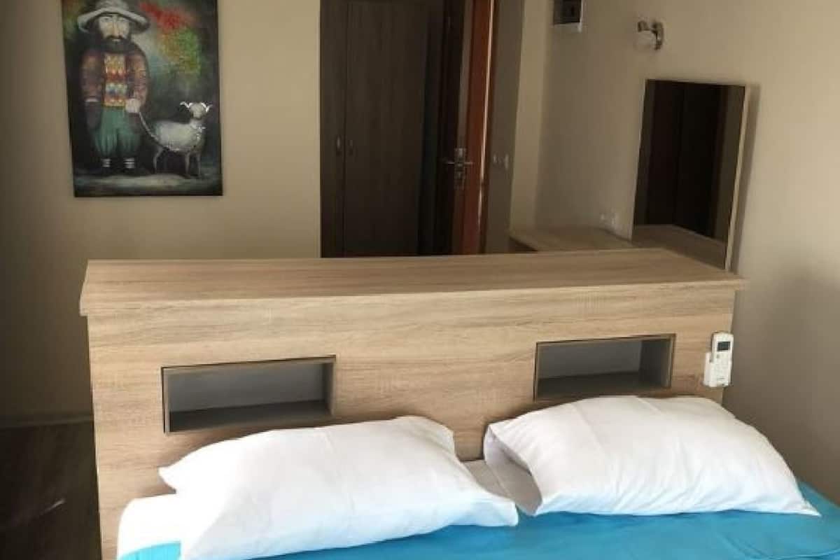 Ein Hotelzimmer mit einem hölzernen Kopfstück, zwei weißen Kissen, einer blauen Tagesdecke und einem Gemälde an der Wand.