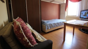 Quarto pequeno com sofá, uma cama de solteiro, mesa de madeira e televisão.