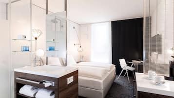 A modern hotel room with a large mirror, a double bed, a bedside table with a lamp, a desk with a chair, and a large window with blinds.