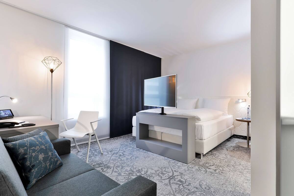 A modern hotel room with a grey sofa, a white bed, a flat-screen TV, a desk with a laptop, and a large window with a black curtain.