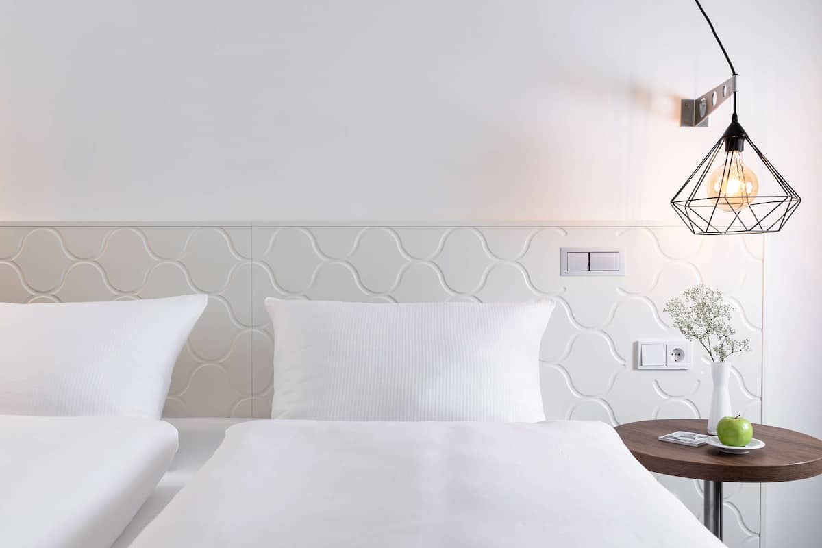 A hotel room with a modern design, featuring a geometric-patterned headboard, a round wooden side table with a vase and an apple, and a unique geometric pendant light.