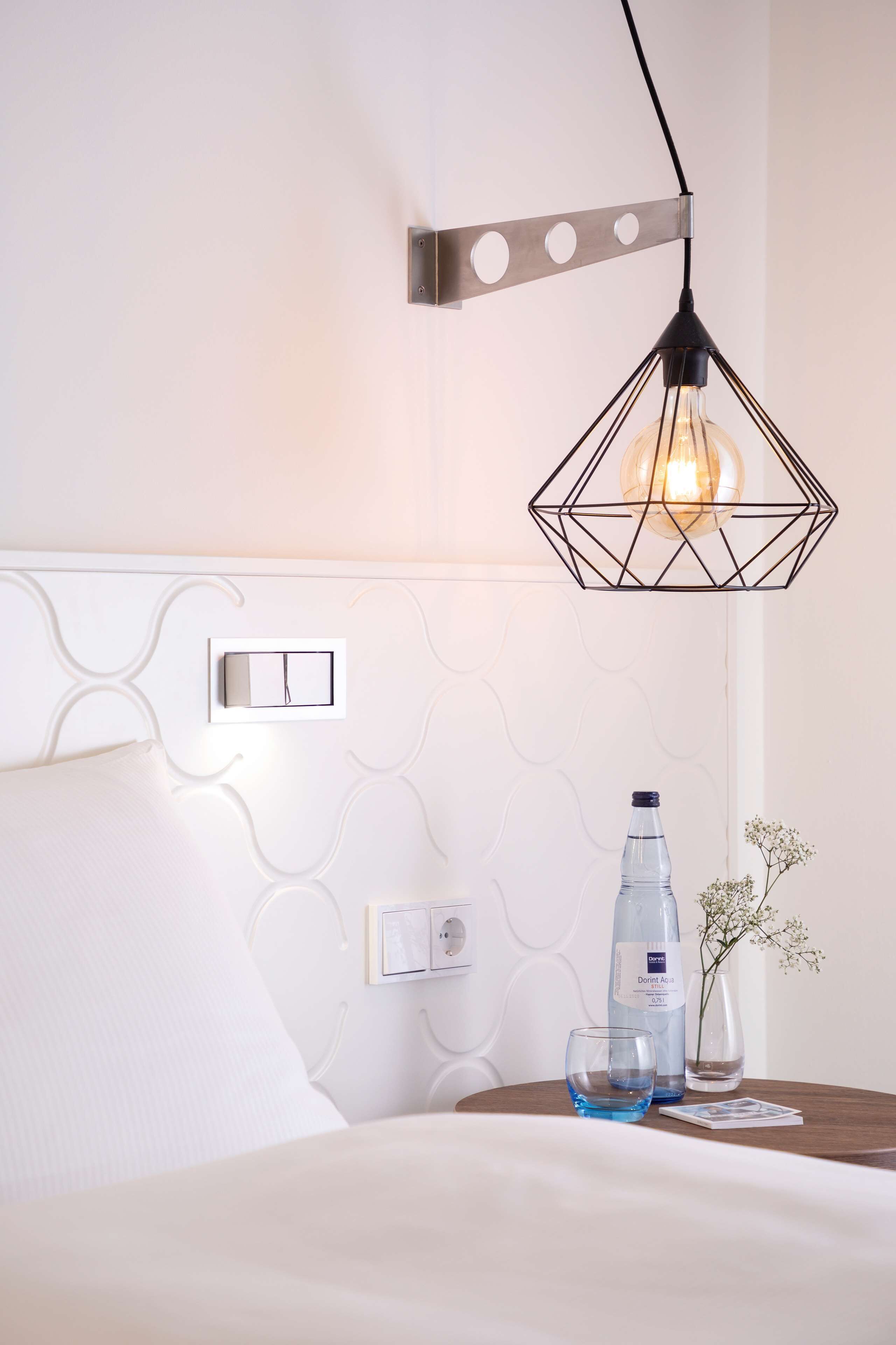 A neatly made bed with a white headboard, a bedside table with a water bottle, a glass, and a small vase with flowers.