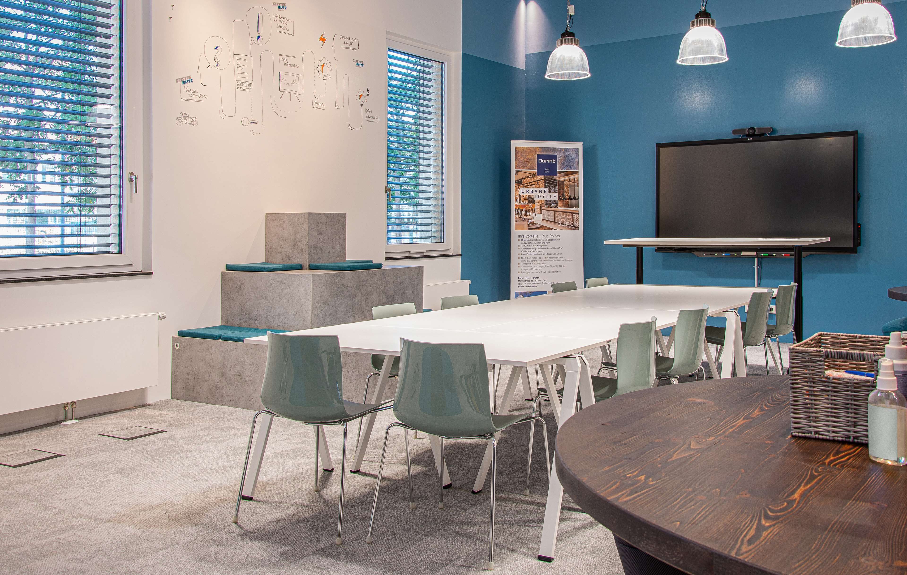 A conference room with a long table, green chairs, a large screen, and a poster on the wall.