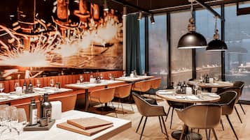 A modern restaurant with a high ceiling, large windows, and a city view. Tables are set with wine glasses, bottles, and menus.