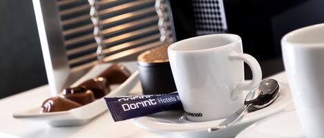 Una preparación de café con una cafetera, dos tazas blancas, una cuchara y un plato pequeño con bombones.