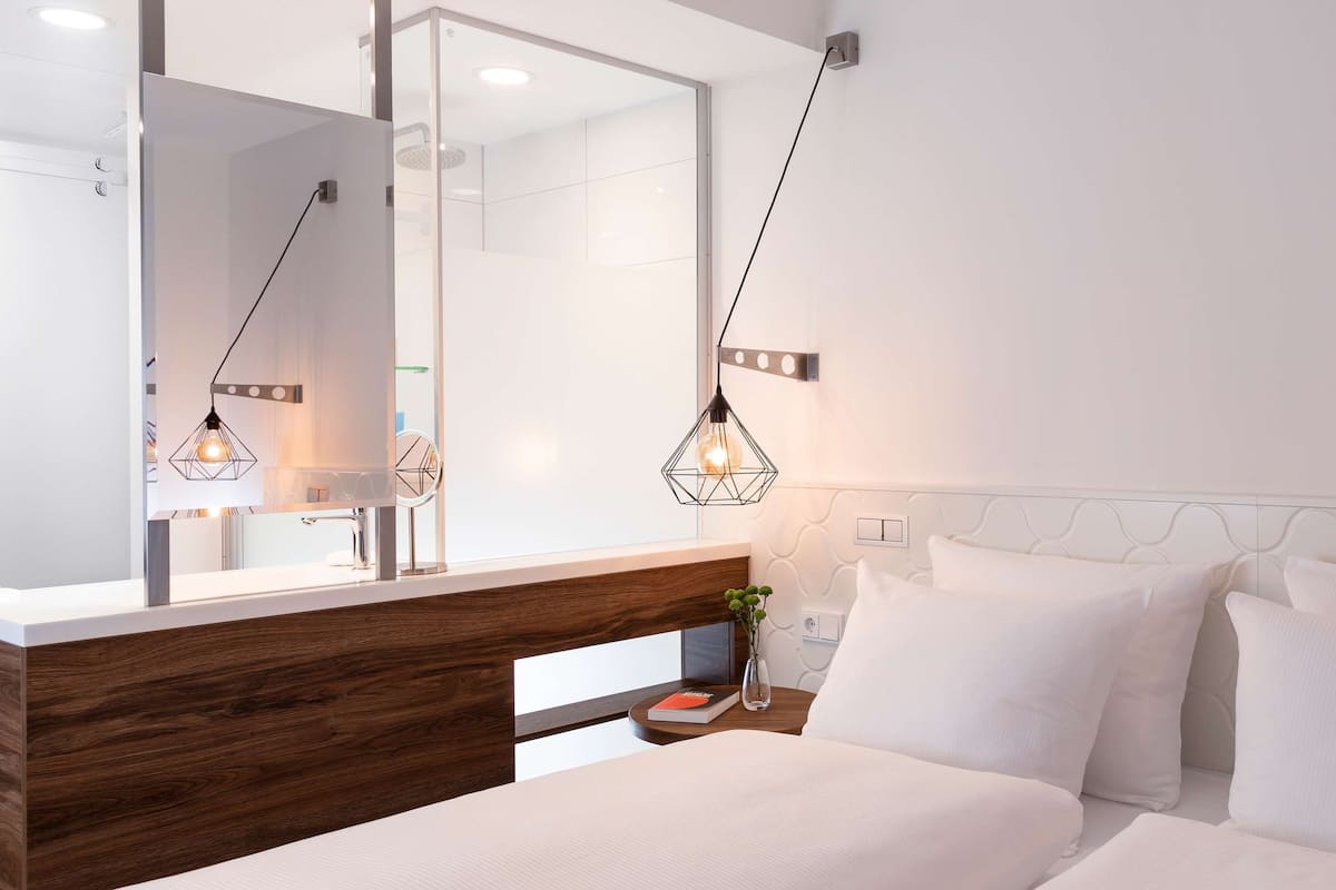 A modern hotel room with a wooden headboard, a large mirror, a bedside table with a book and vase, and a bed with white bedding.