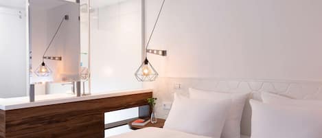 A modern hotel room with a wooden headboard, a large mirror, a bedside table with a book and vase, and a bed with white bedding.