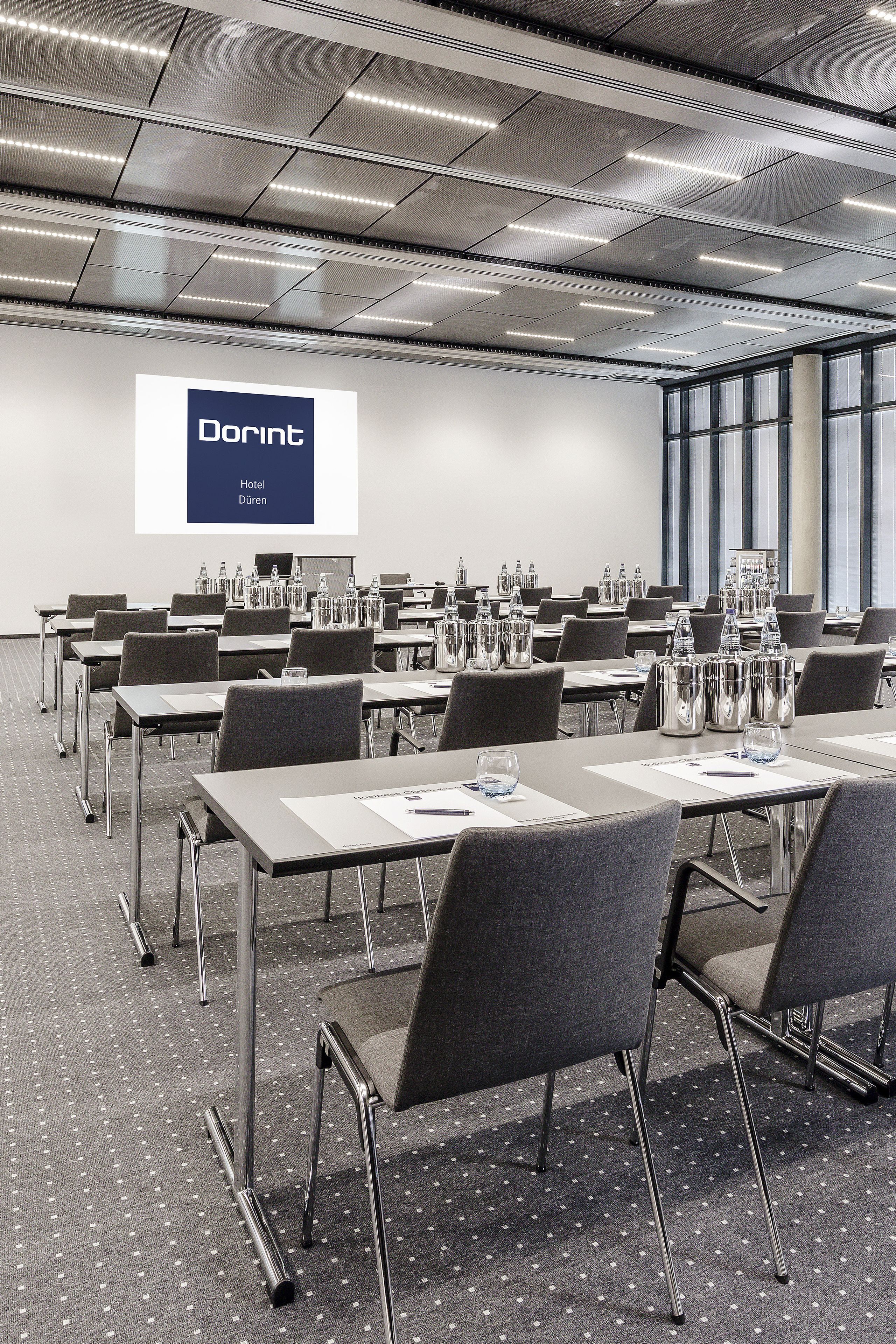Un salón de conferencias con mesas, sillas y una pantalla de proyector que muestra "Dorint Hotel Düren".