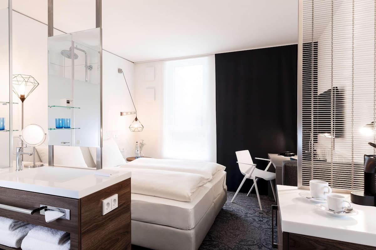 A hotel room with a double bed, a desk, a chair, a large mirror, and a bathroom with a vanity and a shower.