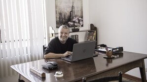 Ein Mann sitzt an einem Schreibtisch mit einem Laptop, einer Maus und einer Tastatur. Auf dem Schreibtisch steht eine Pflanze, im Hintergrund ist ein Fenster mit Jalousien zu sehen.