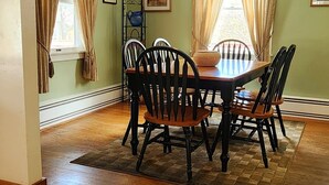 A dining room with a round table, six chairs, and two windows with curtains.