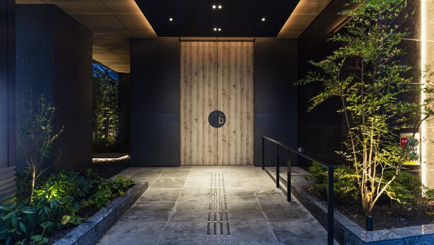 A modern building entrance with a wooden panel wall, a metal railing, and a small tree. - the b asakusa (Tokyo)