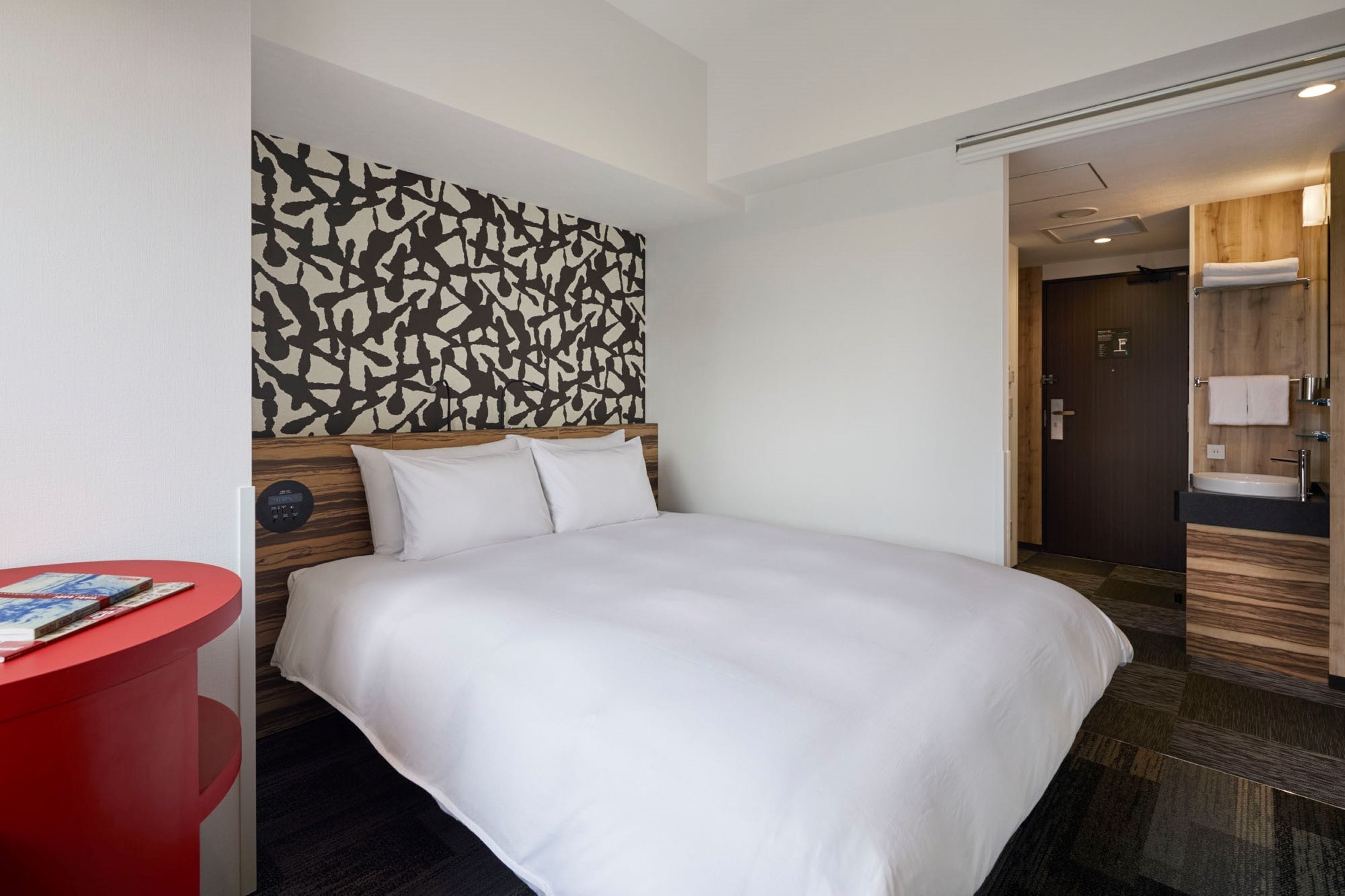 Hotellihuone, jossa on suuri sänky, punainen yöpöytä, moderni abstrakti seinätaideteos ja puinen ovi, joka johtaa kylpyhuoneeseen.