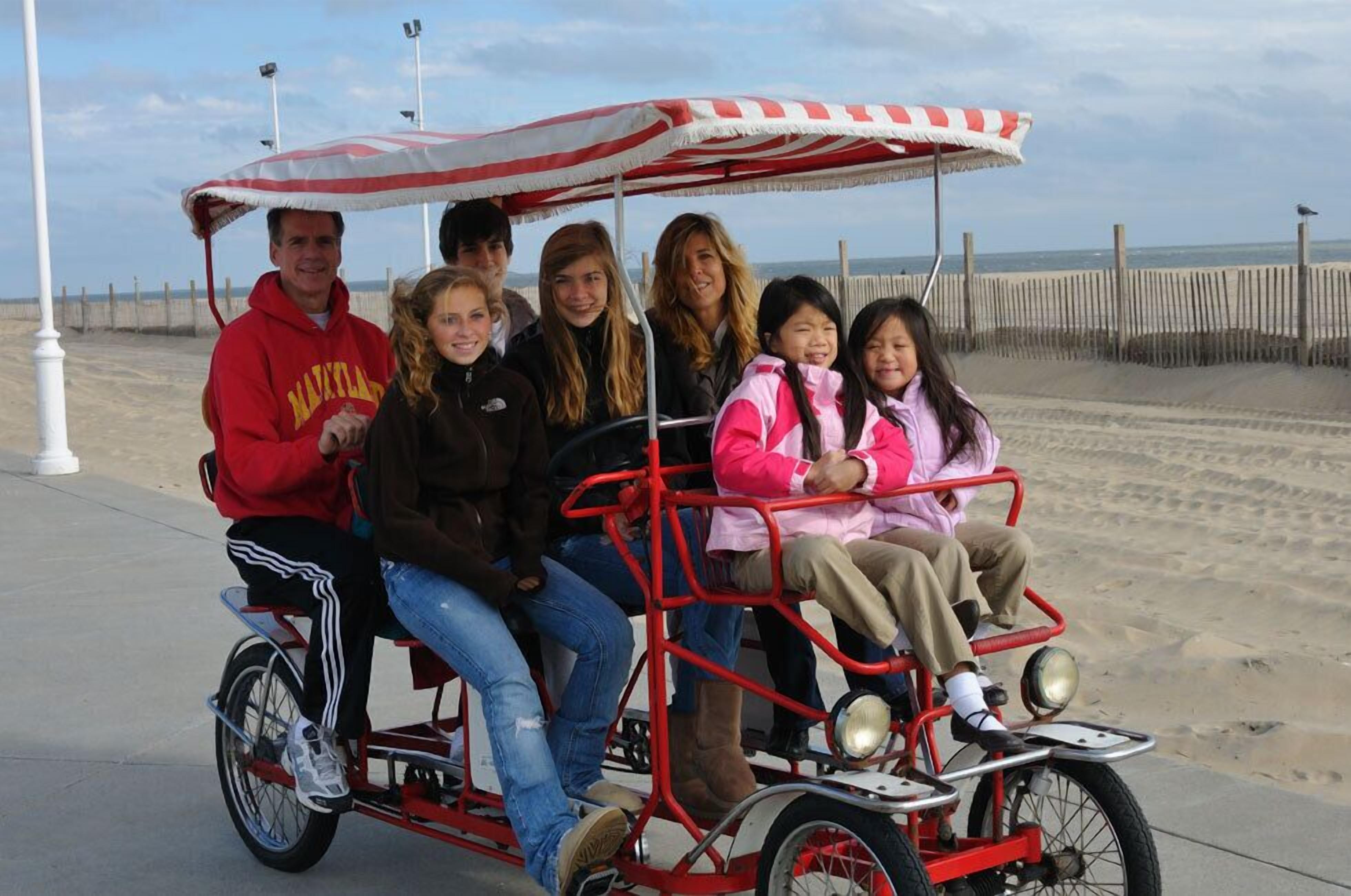 A group of people on a red and white beach buggy with a canopy, parked on a sandy beach with a wooden fence in the background.