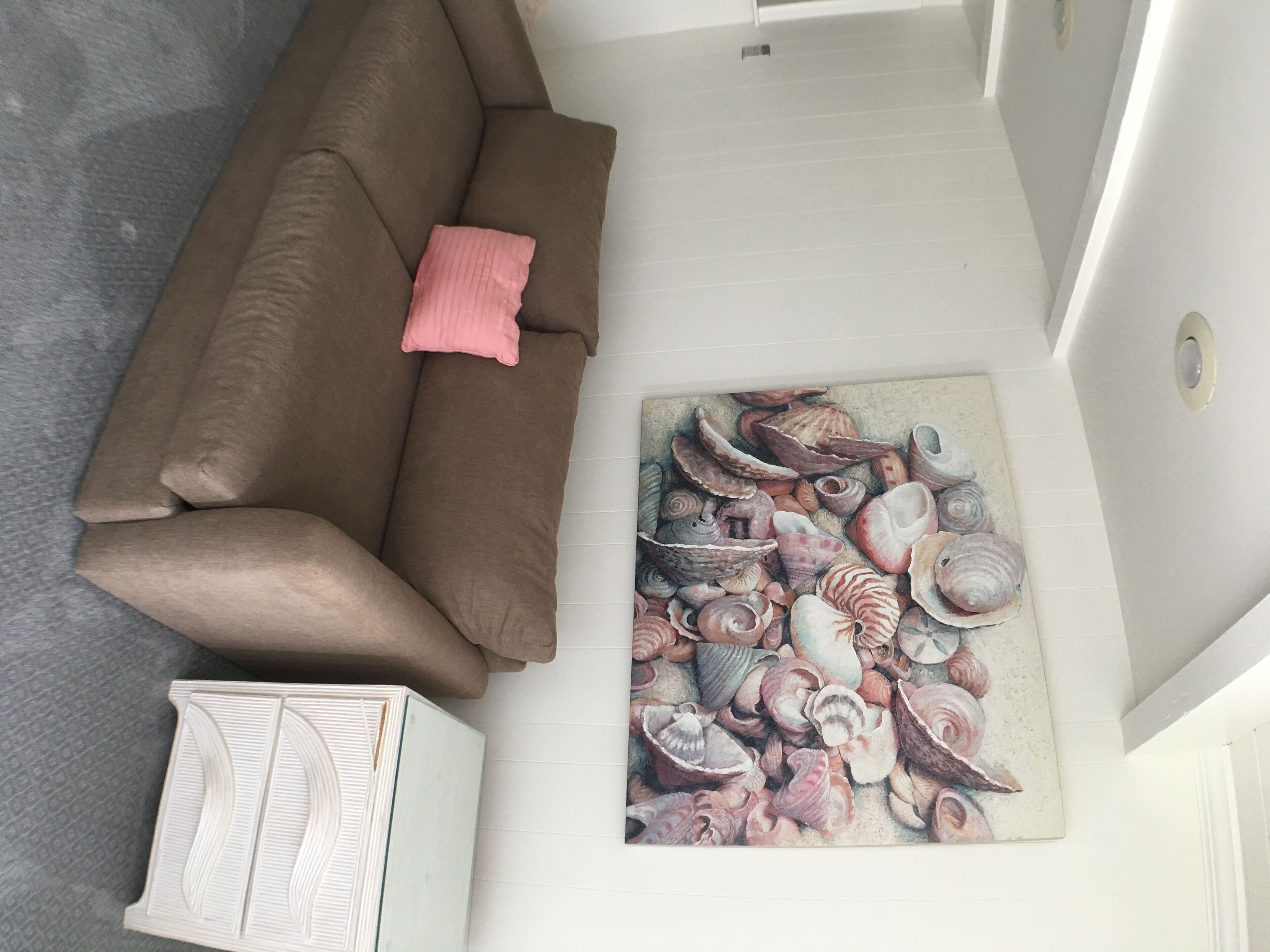 Ein Zimmer mit einem braunen Sofa, einem rosafarbenen Kissen und einem an der Wand hängenden Bild mit Muscheln.