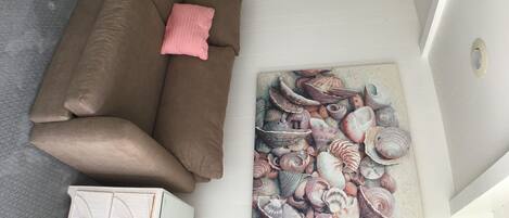 Ein Zimmer mit einem braunen Sofa, einem rosafarbenen Kissen und einem an der Wand hängenden Bild mit Muscheln.