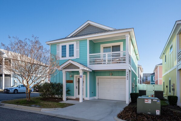 A two-story house with a front garage, a balcony, and a small front yard with a tree and bushes. - Currituck Cottage 3 Bedroom Home at Devonshire Place, Bermuda Bay Amenities! (Kill Devil Hills)