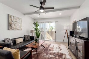 A modern living room with a black leather sofa, a wooden coffee table, a large flat-screen TV, a ceiling fan, and a view of the outdoors through a sliding glass door. - Updated Old Town Pool View Condo | 4 Pools (Scottsdale)