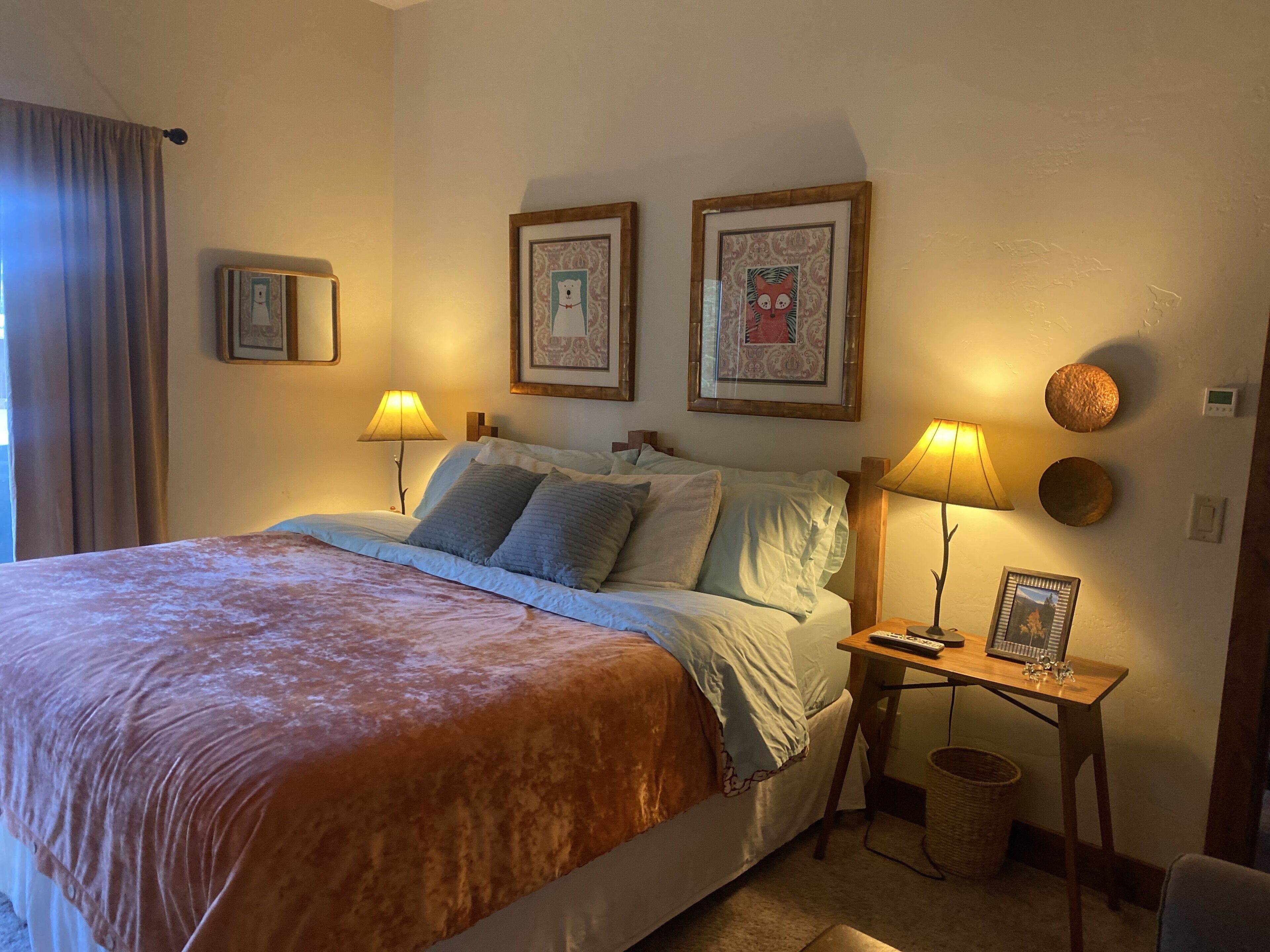 A neatly arranged bedroom with a large bed, two bedside tables with lamps, framed pictures on the wall, and a basket on the floor.