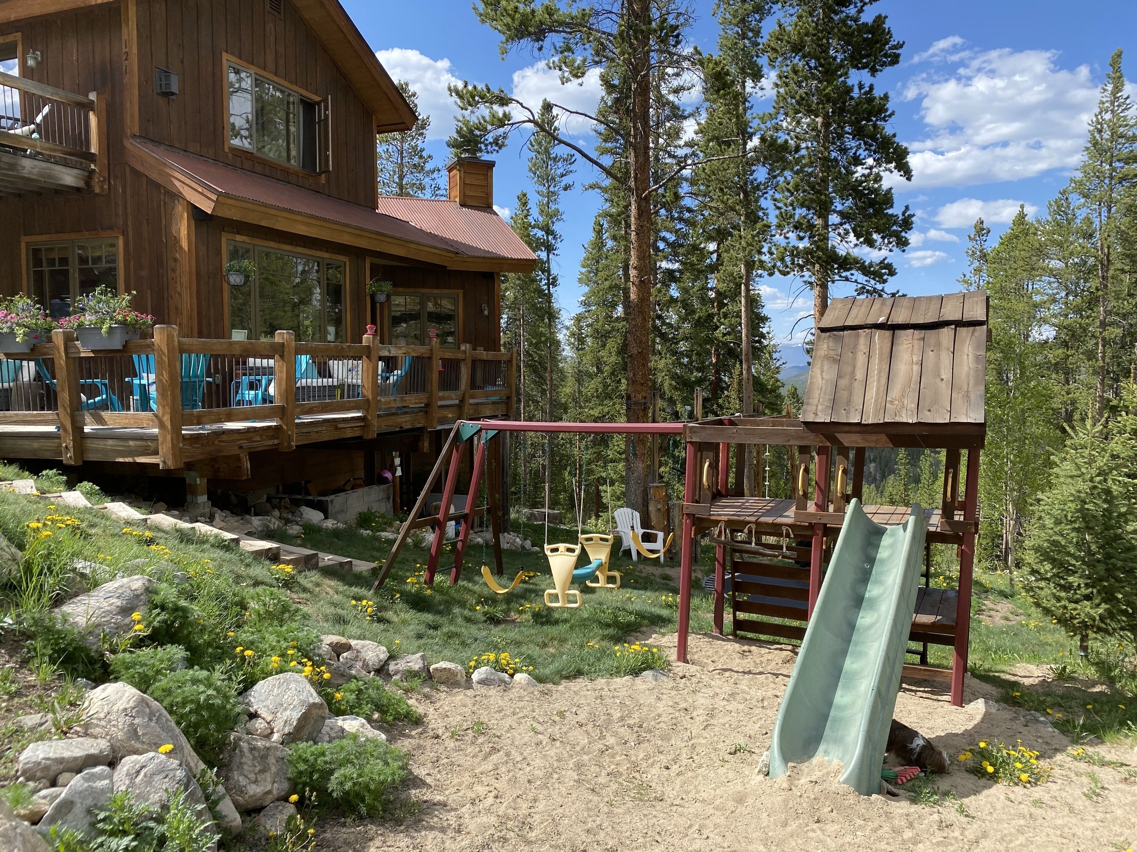 A wooden cabin with a deck, a swing set, and a slide in a forested area.