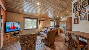 A spacious living room with a television, comfortable seating, a kitchen area, and a dining table.