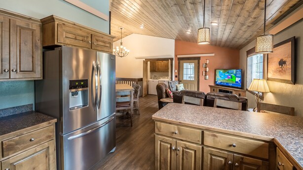 A modern kitchen with a stainless steel refrigerator, wooden cabinets, and a granite countertop. There is a dining area with a table and chairs, and a television mounted on the wall.