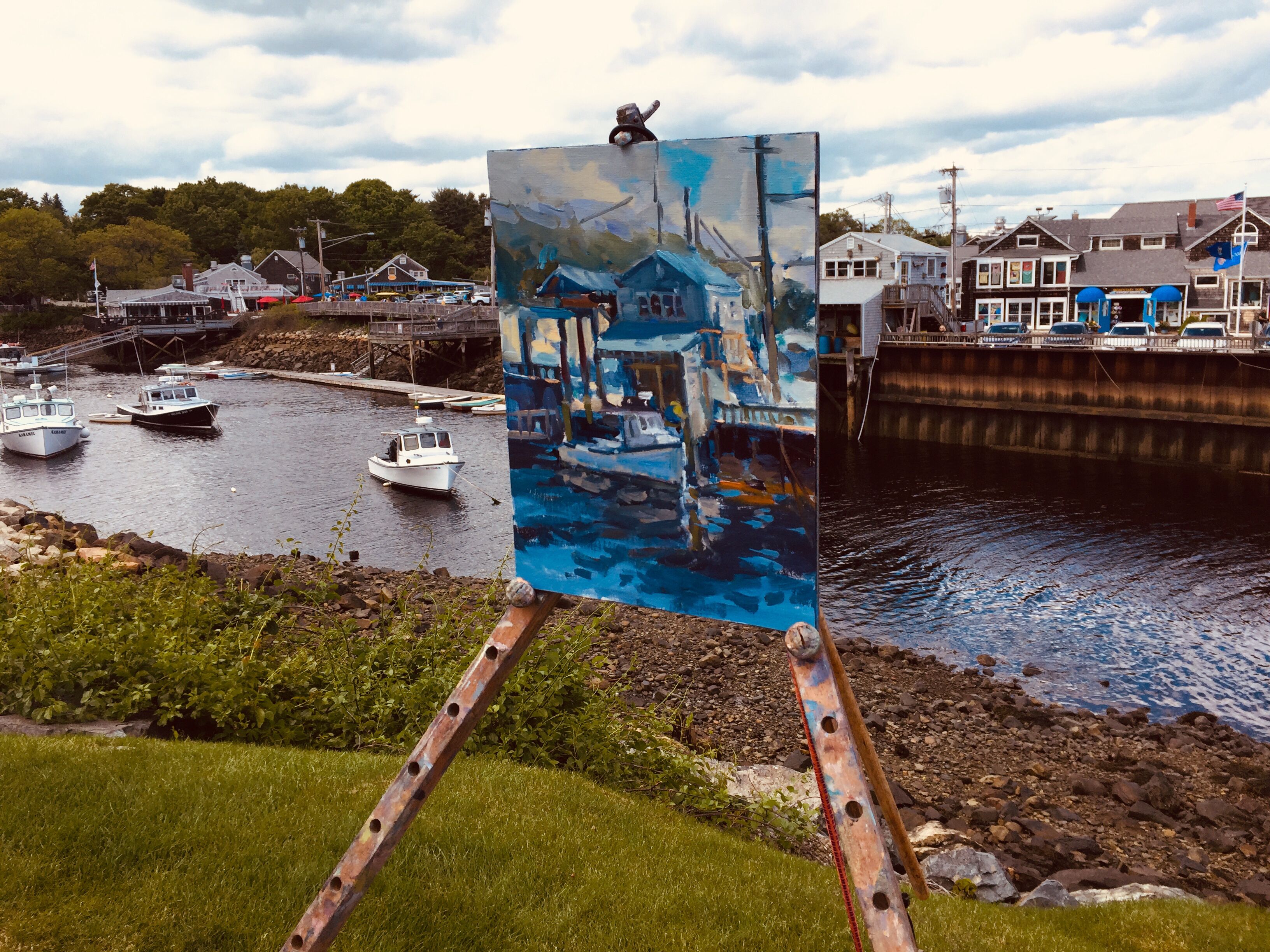 Cena à beira-mar com uma pintura em um cavalete, barcos atracados e edifícios ao fundo.
