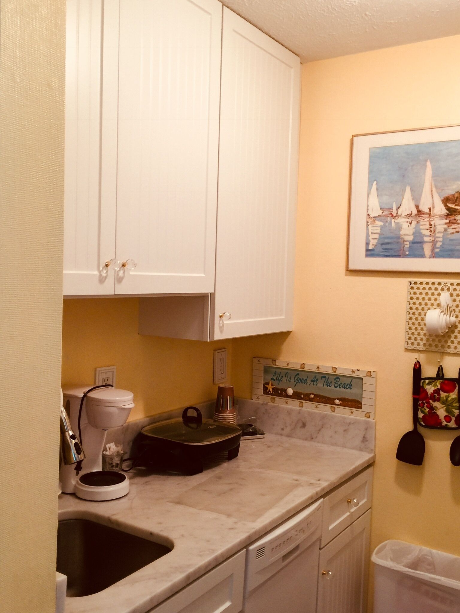 A compact kitchen with white cabinets, a sink, a coffee maker, and a decorative sign on the wall.