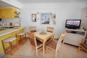 Un coin repas avec une table et des chaises en bois, un petit espace cuisine avec des placards jaunes, un tableau encadré au mur et un téléviseur qui affiche les prévisions météorologiques.