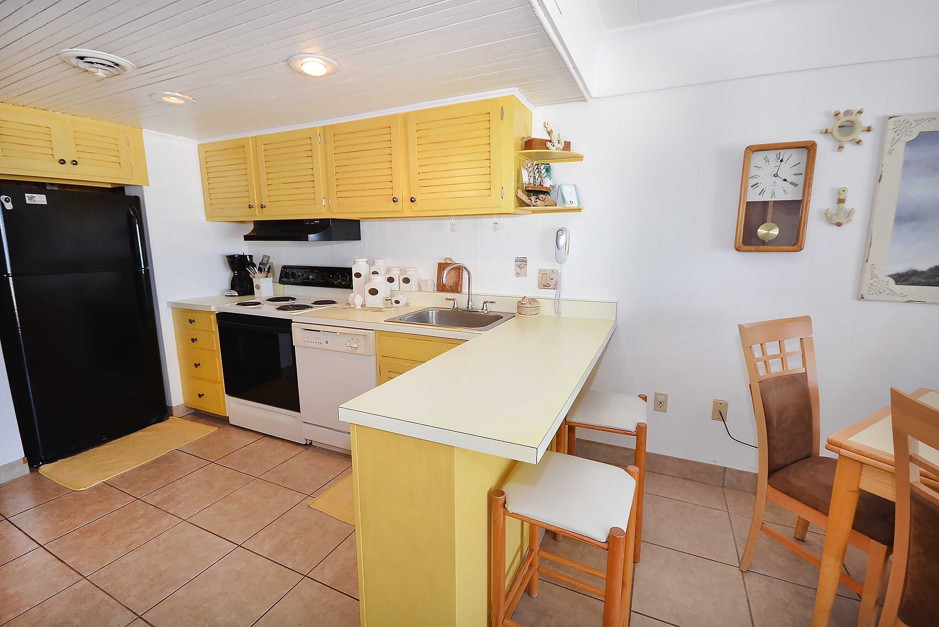 A compact kitchen with yellow cabinets, a black refrigerator, a stove, and a sink. There are two wooden chairs and a small table.